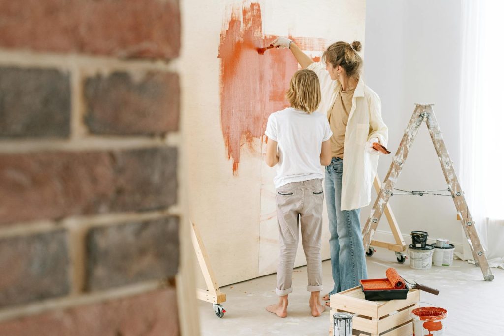 Two people painting a wall red near a brick accent, with a ladder and tools in the background